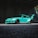 A modified turquoise sports car with a large rear spoiler is parked in an industrial or urban setting, partially shadowed. The car features wide fender flares, low profile tires, and has branding on the side skirts.