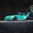 A modified turquoise sports car with a large rear spoiler is parked in an industrial or urban setting, partially shadowed. The car features wide fender flares, low profile tires, and has branding on the side skirts.