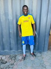 A person is standing on a tiled floor wearing a bright yellow soccer jersey with blue and white trim, blue shorts, white socks, and soccer cleats. The background features a textured, gray metal wall.