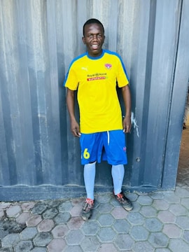 A person is standing on a tiled floor wearing a bright yellow soccer jersey with blue and white trim, blue shorts, white socks, and soccer cleats. The background features a textured, gray metal wall.