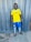 A person is standing on a tiled floor wearing a bright yellow soccer jersey with blue and white trim, blue shorts, white socks, and soccer cleats. The background features a textured, gray metal wall.