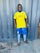 A person is standing on a tiled floor wearing a bright yellow soccer jersey with blue and white trim, blue shorts, white socks, and soccer cleats. The background features a textured, gray metal wall.