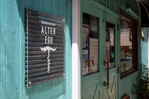 A turquoise-colored storefront with wooden paneling features a modern sign that reads 'Selected wine store ALTER EGO' alongside a graphic of a mustache and wine opener. Posters and notices are visible on the window, reflecting a cozy and inviting atmosphere. The door has a rustic handle, and some greenery is visible nearby.