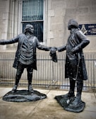 Two bronze statues depict historical figures in colonial attire. They stand facing each other, extending a handshake, with a building and a Masonic Temple plaque in the background. Snowflakes are visible, suggesting a cold atmosphere.
