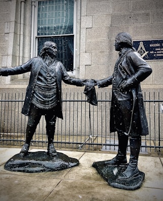 Two bronze statues depict historical figures in colonial attire. They stand facing each other, extending a handshake, with a building and a Masonic Temple plaque in the background. Snowflakes are visible, suggesting a cold atmosphere.
