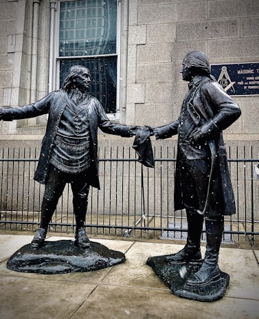 Two bronze statues depict historical figures in colonial attire. They stand facing each other, extending a handshake, with a building and a Masonic Temple plaque in the background. Snowflakes are visible, suggesting a cold atmosphere.