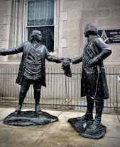 Two bronze statues depict historical figures in colonial attire. They stand facing each other, extending a handshake, with a building and a Masonic Temple plaque in the background. Snowflakes are visible, suggesting a cold atmosphere.