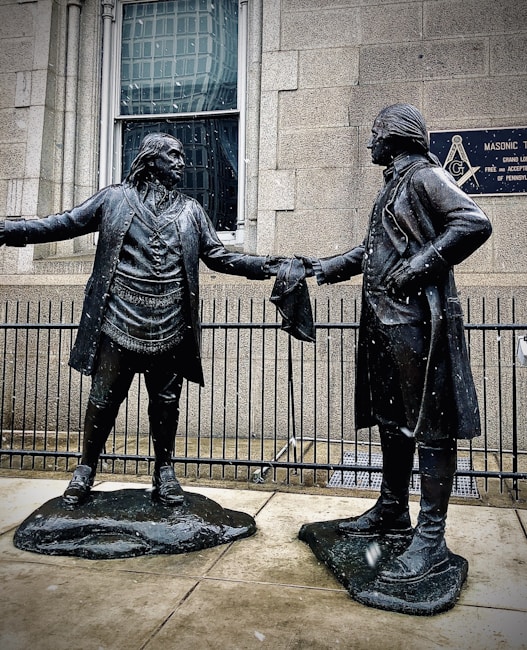 Two bronze statues depict historical figures in colonial attire. They stand facing each other, extending a handshake, with a building and a Masonic Temple plaque in the background. Snowflakes are visible, suggesting a cold atmosphere.