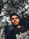 A young man is standing outdoors, looking directly at the camera with a confident expression. He is wearing a dark t-shirt that reads 'Believe in Yourself' in white script. The background consists of blurred tree branches and leaves, creating a natural setting with a focus on the person in the foreground.