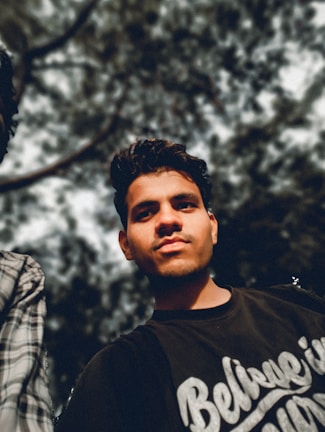A young man is standing outdoors, looking directly at the camera with a confident expression. He is wearing a dark t-shirt that reads 'Believe in Yourself' in white script. The background consists of blurred tree branches and leaves, creating a natural setting with a focus on the person in the foreground.