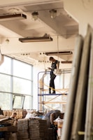 Workers are assembling or disassembling materials in a large industrial or warehouse space with tall windows. One person stands on a scaffold, seemingly adjusting a light fixture or ceiling component. Another individual is bent over working with materials stacked on a table or platform.