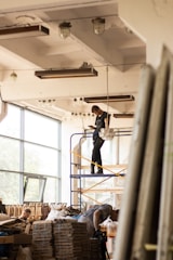 Workers are assembling or disassembling materials in a large industrial or warehouse space with tall windows. One person stands on a scaffold, seemingly adjusting a light fixture or ceiling component. Another individual is bent over working with materials stacked on a table or platform.