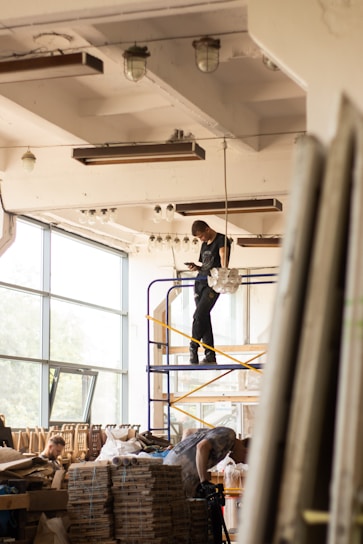 Workers are assembling or disassembling materials in a large industrial or warehouse space with tall windows. One person stands on a scaffold, seemingly adjusting a light fixture or ceiling component. Another individual is bent over working with materials stacked on a table or platform.
