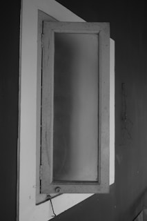 A monochrome image of a slightly open window with frosted glass, set against a dark wall. The window frame is rectangular with visible hinges, and there is a faint reflection or pattern on the glass. The contrast between the light-colored window frame and the darker surrounding wall is prominent.