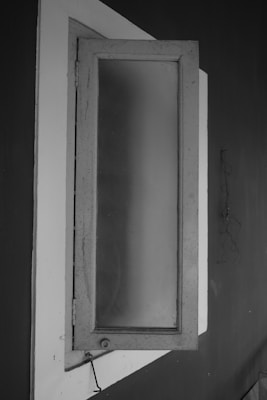 A monochrome image of a slightly open window with frosted glass, set against a dark wall. The window frame is rectangular with visible hinges, and there is a faint reflection or pattern on the glass. The contrast between the light-colored window frame and the darker surrounding wall is prominent.
