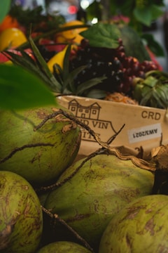 Several green coconuts are placed in the foreground, with their rough texture and natural patterns visible. Behind them, a wooden box features a design and some text, partially obscured. In the background, a variety of colorful fruits and vegetables add visual interest, including red and yellow tones. The overall composition feels fresh and vibrant, with some leaves and branches adding to the natural setting.