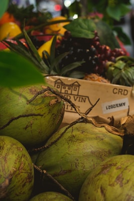 Several green coconuts are placed in the foreground, with their rough texture and natural patterns visible. Behind them, a wooden box features a design and some text, partially obscured. In the background, a variety of colorful fruits and vegetables add visual interest, including red and yellow tones. The overall composition feels fresh and vibrant, with some leaves and branches adding to the natural setting.