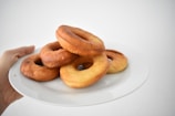 A stack of golden-brown doughnuts is placed on a white plate, held up by a hand against a plain white background. The doughnuts are freshly baked with varying tones of brown and yellow.