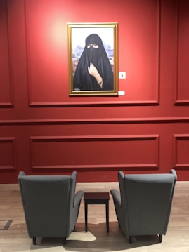 A gallery setting with a large portrait of a woman wearing a black niqab. The portrait is framed in gold and hangs on a rich red wall. In front of the painting are two grey armchairs with a small dark wooden table between them, placed on a light wooden floor.
