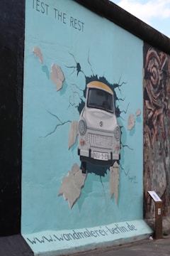 A mural on the Berlin Wall depicting a white car breaking through the wall, surrounded by cracks and debris. The car has a license plate reading 'NOV-9-89.' The wall is painted a light blue color with visible text 'Test the Rest' at the top. There's a small sign or plaque next to the mural.