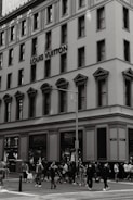 A large, multi-story building features classic architecture with arched windows and ornate moldings. The building prominently displays the Louis Vuitton name. A busy street intersection is populated with pedestrians, some of whom are wearing masks and carrying shopping bags.
