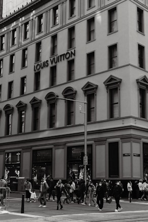 A large, multi-story building features classic architecture with arched windows and ornate moldings. The building prominently displays the Louis Vuitton name. A busy street intersection is populated with pedestrians, some of whom are wearing masks and carrying shopping bags.