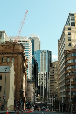 Tall modern skyscrapers and older historical buildings line a busy city street. A crane is visible against a clear blue sky, indicating ongoing construction. Traffic lights and road cones suggest urban development or maintenance work.
