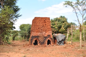 A tall stack of red clay bricks are arranged in a pyramidal structure surrounded by lush greenery. Two arch-shaped openings are visible at the base, resembling a kiln or oven. A black tarp partially covers the right side of the structure. The ground is a reddish-brown earth, and trees with dense foliage frame the scene.