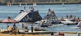 A lively outdoor scene featuring an inflatable water park with people actively engaging in climbing and water activities. The park is set on a lake or river with several sailboats in the background. Nearby, a group of people are relaxing under a tent, and others are on a dock with kayaks.