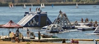 A lively outdoor scene featuring an inflatable water park with people actively engaging in climbing and water activities. The park is set on a lake or river with several sailboats in the background. Nearby, a group of people are relaxing under a tent, and others are on a dock with kayaks.