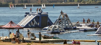 A lively outdoor scene featuring an inflatable water park with people actively engaging in climbing and water activities. The park is set on a lake or river with several sailboats in the background. Nearby, a group of people are relaxing under a tent, and others are on a dock with kayaks.