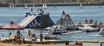 A lively outdoor scene featuring an inflatable water park with people actively engaging in climbing and water activities. The park is set on a lake or river with several sailboats in the background. Nearby, a group of people are relaxing under a tent, and others are on a dock with kayaks.