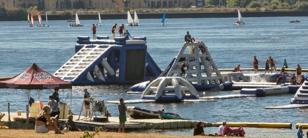 A lively outdoor scene featuring an inflatable water park with people actively engaging in climbing and water activities. The park is set on a lake or river with several sailboats in the background. Nearby, a group of people are relaxing under a tent, and others are on a dock with kayaks.