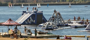 A lively outdoor scene featuring an inflatable water park with people actively engaging in climbing and water activities. The park is set on a lake or river with several sailboats in the background. Nearby, a group of people are relaxing under a tent, and others are on a dock with kayaks.