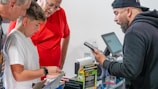 Several people are gathered around a checkout counter. A man wearing a cap and hoodie holds a smartphone, engaging with a boy in a white t-shirt who is closely examining items, possibly trading cards, on the counter. An older man in a red shirt is also observing the scene, looking down at the items.