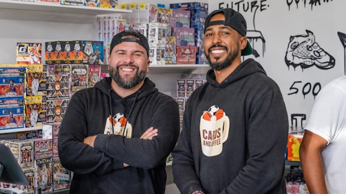 Two smiling men stand inside a shop selling sports cards. They both wear black hoodies with a logo that says 'Cans and Coffee'. Numerous boxes of trading cards are neatly stacked on shelves behind them. The shop's walls feature art and doodles, including a detailed drawing of a sneaker.