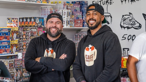 Two smiling men stand inside a shop selling sports cards. They both wear black hoodies with a logo that says 'Cans and Coffee'. Numerous boxes of trading cards are neatly stacked on shelves behind them. The shop's walls feature art and doodles, including a detailed drawing of a sneaker.