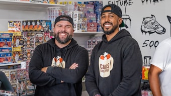 Two smiling men stand inside a shop selling sports cards. They both wear black hoodies with a logo that says 'Cans and Coffee'. Numerous boxes of trading cards are neatly stacked on shelves behind them. The shop's walls feature art and doodles, including a detailed drawing of a sneaker.