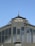 An ornate metal structure displays the words 'Ferrovie Nord Milano' at the top, with 'ARRIVI' below. The architectural style includes rivets and decorative elements, framed against a clear blue sky.