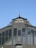 An ornate metal structure displays the words 'Ferrovie Nord Milano' at the top, with 'ARRIVI' below. The architectural style includes rivets and decorative elements, framed against a clear blue sky.
