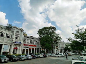 A bustling urban scene with colonial-style white buildings featuring large windows and signs for various shops. In the foreground, a row of parked cars lines the street, with a few people walking by. Trees with lush green foliage are interspersed among the buildings, and the sky is partially cloudy with patches of blue.