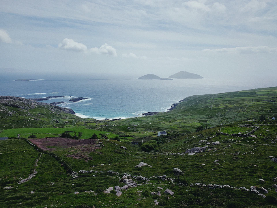 The image depicts a picturesque coastal landscape with two islands visible in the distance, one larger and more prominent island on the left side and another smaller island towards the right. The scene is dominated by a vast expanse of water that stretches out to meet the sky at the horizon line. The land is lush and green, with patches of grass interspersed among rocks and boulders scattered across the landscape. The image does not contain any discernible text or human-made objects, giving it an untouched, serene quality.