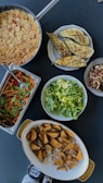 An assortment of dishes on a dark tabletop includes a pan of what appears to be couscous with vegetables, grilled eggplants on a plate, a serving tray with a colorful salad of greens, carrots, and tomatoes, a large bowl of fresh green lettuce, a dish with roasted potato wedges, and a small bowl of mixed mushrooms. Two small jars with labels can be seen at the bottom edge of the image.