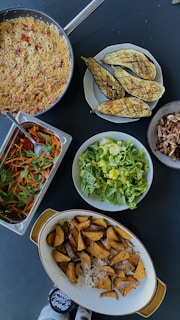 An assortment of dishes on a dark tabletop includes a pan of what appears to be couscous with vegetables, grilled eggplants on a plate, a serving tray with a colorful salad of greens, carrots, and tomatoes, a large bowl of fresh green lettuce, a dish with roasted potato wedges, and a small bowl of mixed mushrooms. Two small jars with labels can be seen at the bottom edge of the image.