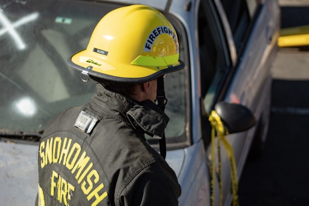 A firefighter, wearing a yellow helmet and a black fire jacket with the words 'SNOHOMISH FIRE' on the back, is standing next to a silver car. The car has yellow caution tape around its side mirror and appears to have been involved in an incident.