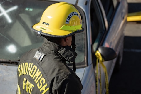 A firefighter, wearing a yellow helmet and a black fire jacket with the words 'SNOHOMISH FIRE' on the back, is standing next to a silver car. The car has yellow caution tape around its side mirror and appears to have been involved in an incident.