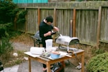 A person is preparing food outdoors using a small, portable oven on a wooden table. The table is cluttered with various kitchen items such as a bowl of chopped vegetables, paper towels, and cooking utensils. The setting is a backyard, featuring dry grass, a wooden fence, and surrounding greenery.