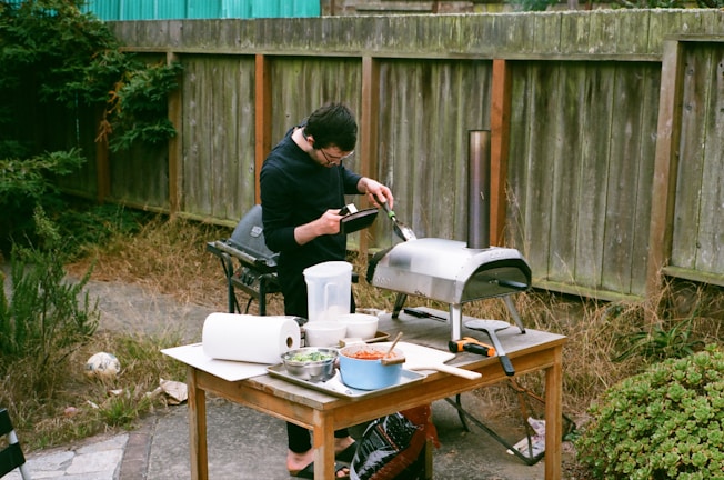A person is preparing food outdoors using a small, portable oven on a wooden table. The table is cluttered with various kitchen items such as a bowl of chopped vegetables, paper towels, and cooking utensils. The setting is a backyard, featuring dry grass, a wooden fence, and surrounding greenery.