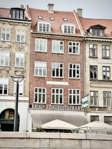 A row of historic European-style buildings with brick facades, featuring large windows and red-tiled roofs. The central building hosts a restaurant called 'Krogs Fiskerestaurant', indicated by signage above the entrance. The lower area includes an outdoor seating area with umbrellas. A modern streetlamp is visible in the foreground.