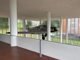 A vintage airplane is suspended within a modern architectural space, featuring large windows and white pillars. The plane has a grey body with black and white markings, and a wooden propeller at the front. The background shows a lush green landscape through glass.